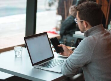 man looking at a smart phone with a laptop on the desk