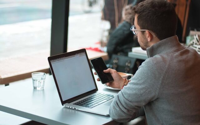 man looking at a smart phone with a laptop on the desk